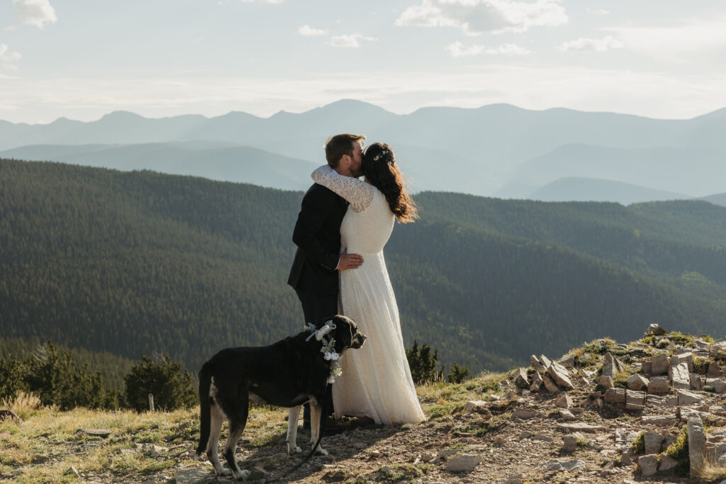 A couple embraces in their wedding attire at a mountain overlook on a sunny day.