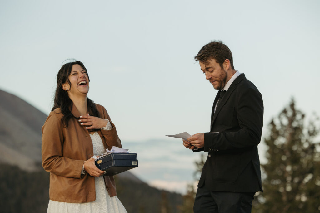 A bride laughs while her husband reads a letter from family members during their elopement at a mountain overlook.