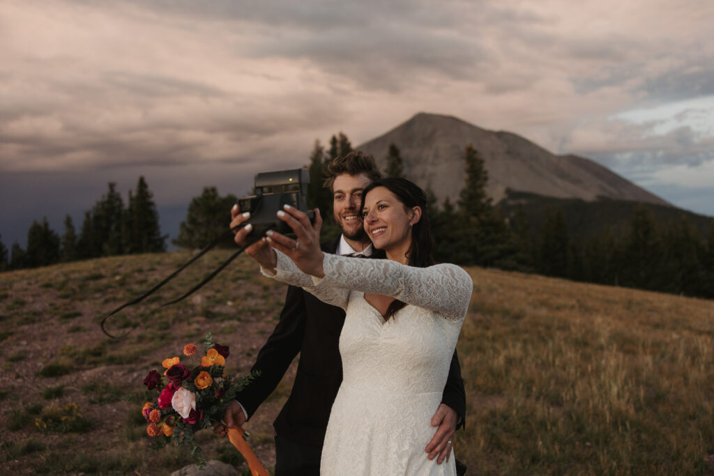 A couple takes a selfie with a Polaroid camera at sunset in front of a mountain on their wedding day.