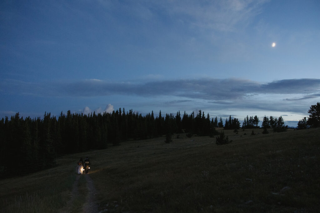 A couple hikes on a trail at blue hour with the moon above them after eloping in the mountains.